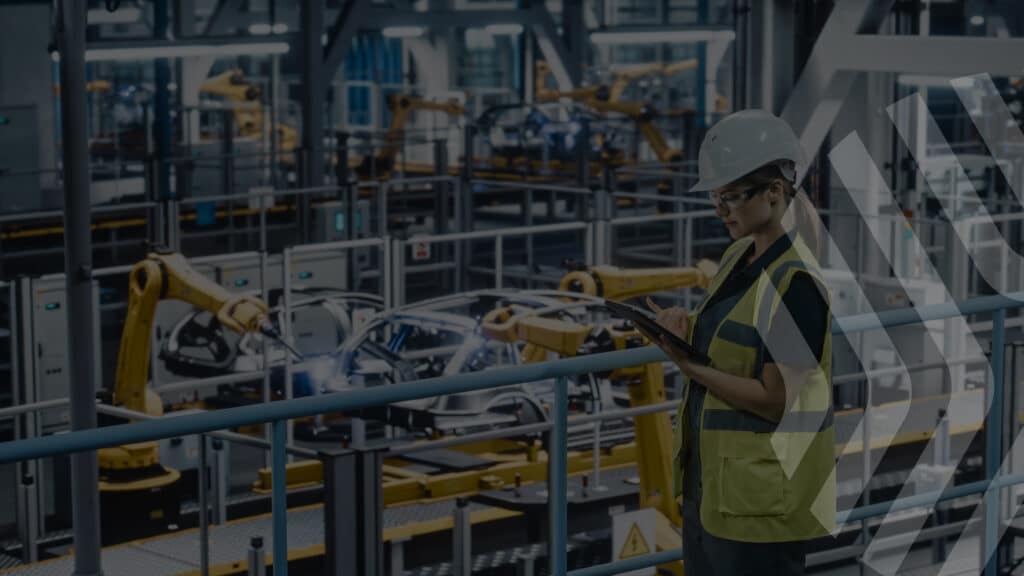 A woman wearing a hard hat, safety glasses, and a yellow vest uses a tablet while standing in a modern manufacturing facility with robotic arms and car frames in the background.
