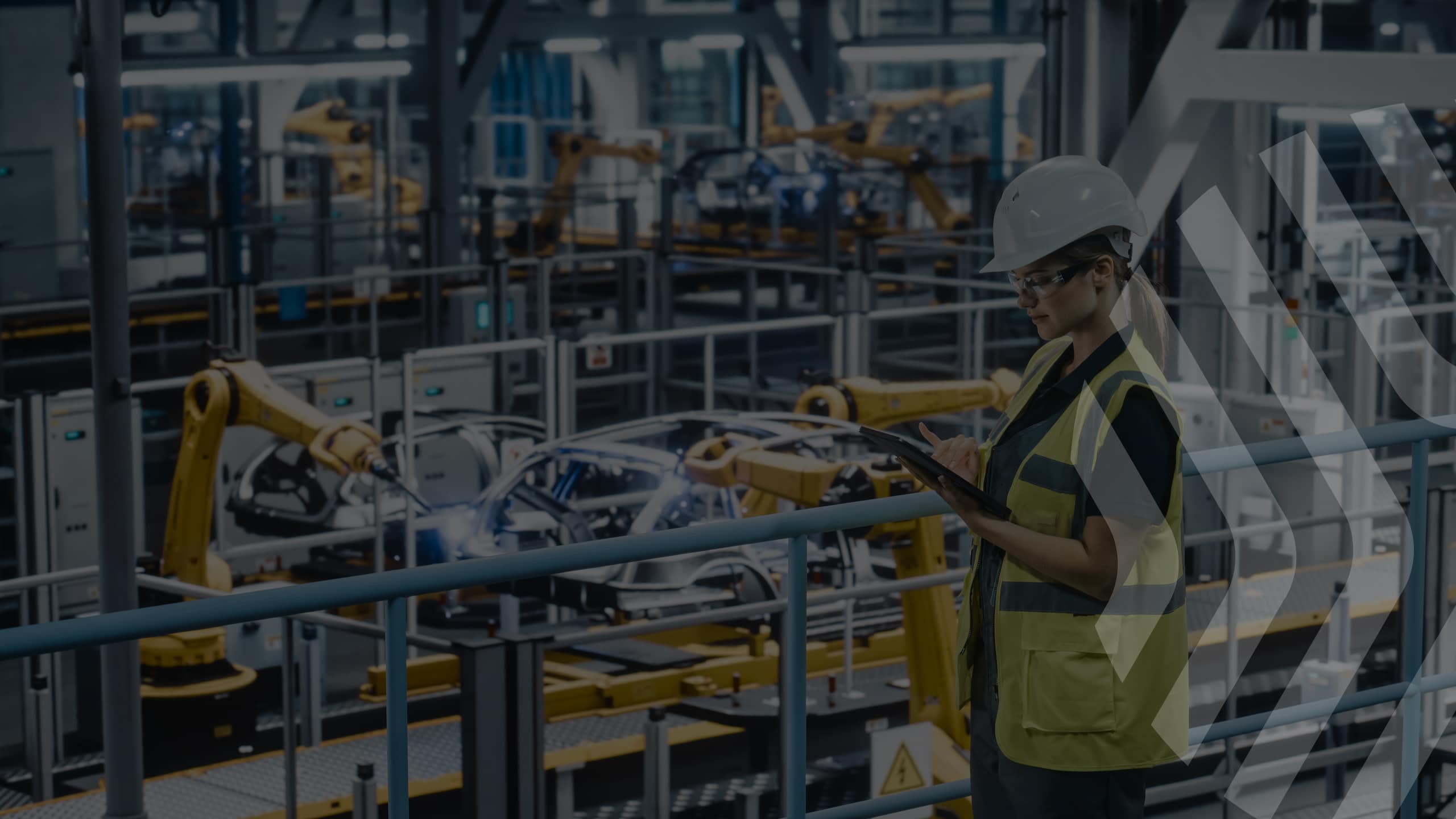 A woman wearing a hard hat, safety glasses, and a yellow vest uses a tablet while standing in a modern manufacturing facility with robotic arms and car frames in the background.
