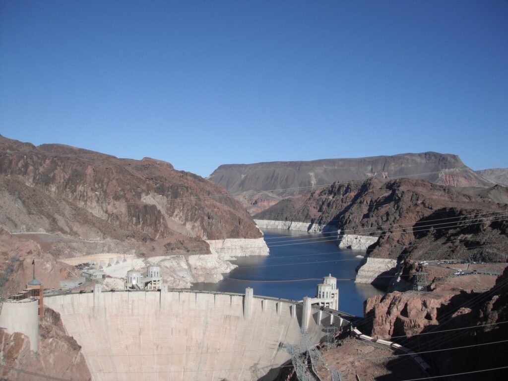 A large concrete dam, built as part of major dam projects, spans a river between rocky, brown mountains with a blue sky above. The reservoir behind the dam has a visible white bathtub ring along the shoreline.