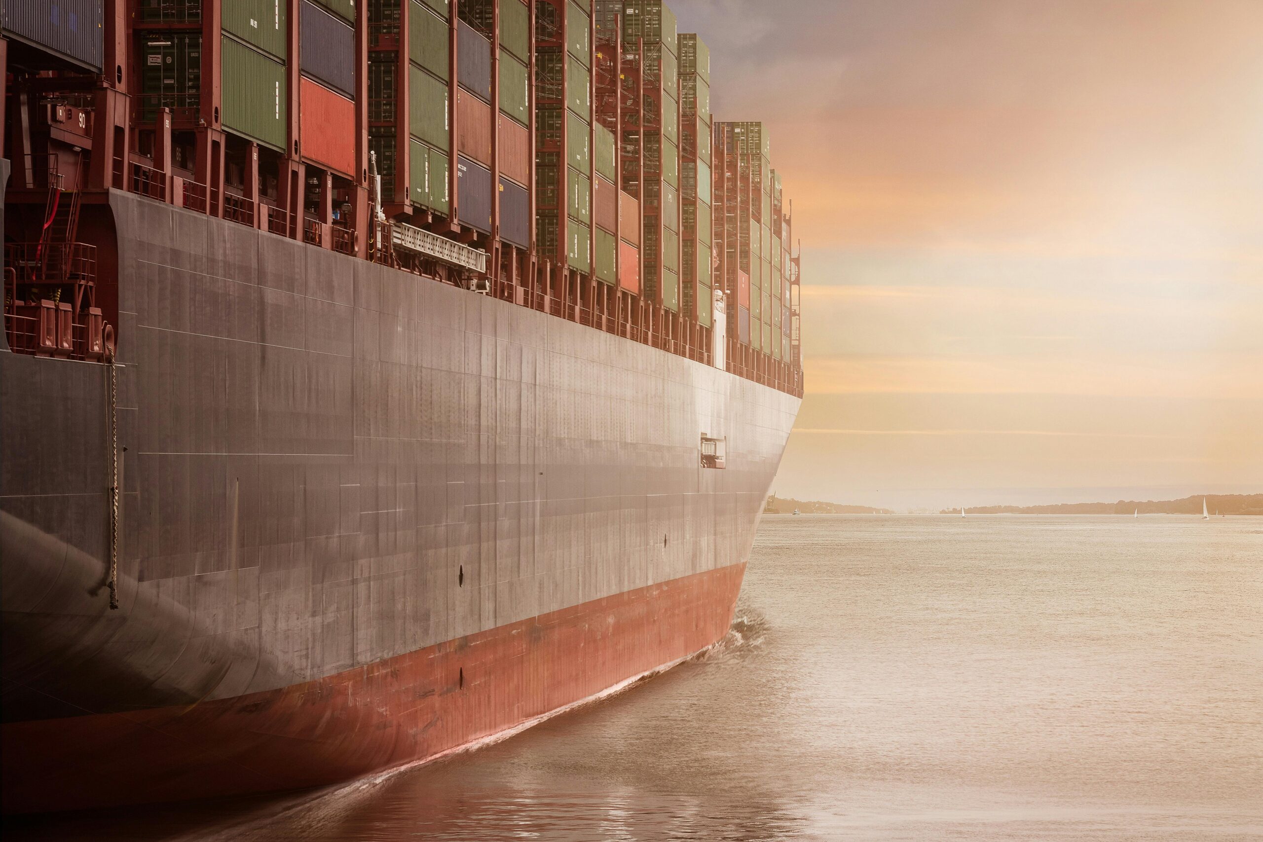 A large cargo ship loaded with stacked shipping containers sails on calm water at sunset, with a hazy sky and distant shoreline in the background—highlighting the importance of safety to prevent transportation accidents at sea.