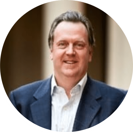 A man with short brown hair wearing a dark suit jacket and white shirt smiles at the camera, standing in front of a blurred background with columns.