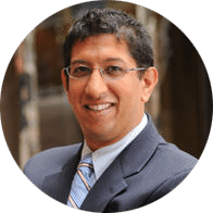 A man with dark hair and glasses, wearing a dark suit, light blue shirt, and striped tie, smiling while posing for a professional headshot in an indoor setting.
