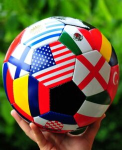 A hand holds a soccer ball decorated with various national flags, including those of the USA, Mexico, Finland, and Turkey. Against blurred greenery, the image reflects excitement for World Cup 2026 safety and celebration.
